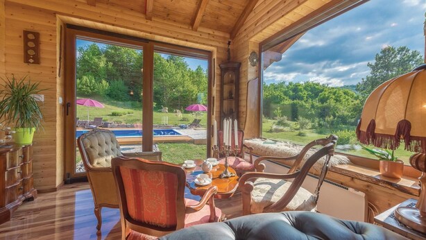 A cozy room with a wooden ceiling, large windows, a dining table with chairs, and a view of a swimming pool and greenery outside. - Lovely home in Gospic (Gospic)