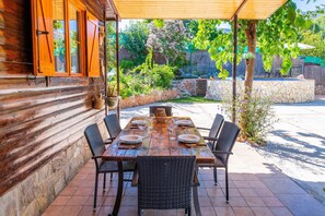 A wooden table set for dining with wicker chairs, a stone wall, and a garden with various plants and a wooden fence in the background. - Cubo's Finca La Rosa del Zangano / Rural / pool / barbecue / WIFI (Alhaurín el Grande)