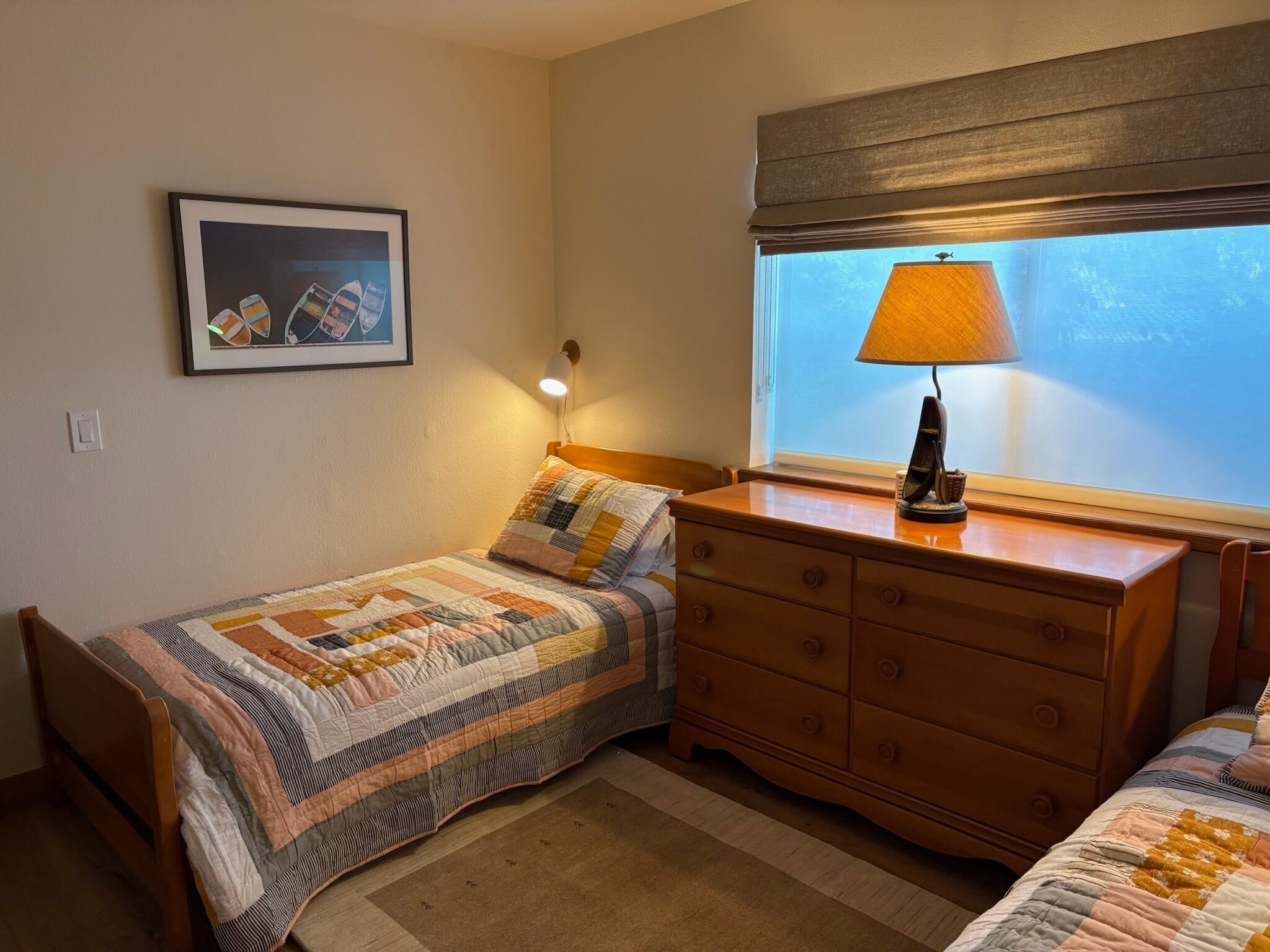 Quarto com duas camas, cômoda, abajur, quadro na parede e janela com vista.