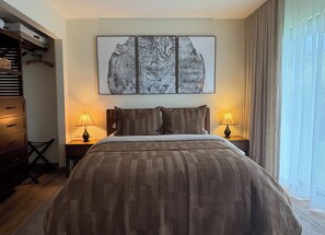 Een hotelkamer met een bed, twee bedlampen, een groot kunstwerk in een lijst boven het bed, en een raam met gordijnen.