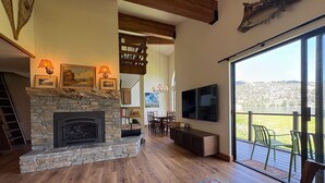 A spacious living room with a stone fireplace, wooden flooring, a large TV, and a balcony with a view of the countryside. - Lakefront retreat with big views | Freshly renovated (June Lake)