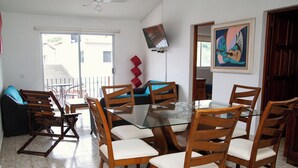 A dining area with a glass table surrounded by wooden chairs with white cushions, a sofa, and a television mounted on the wall. - Casa Bahía Department 1 (Centro)