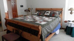 A bedroom with a wooden bed, a quilt, a chair, a bedside table, and a window with blinds. - Hideaway #103, views of Carson Peak (June Lake)