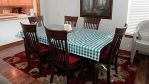 A dining area with a table set for six, wooden chairs, a checkered tablecloth, a painting on the wall, and a kitchen in the background. - Hideaway #103, views of Carson Peak (June Lake)