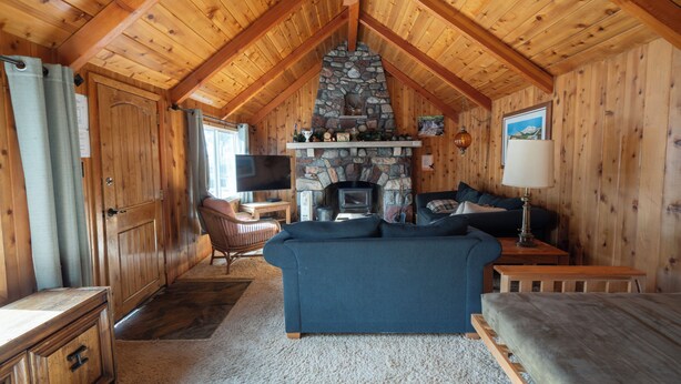 A cozy cabin interior with a stone fireplace, wooden ceiling beams, a sofa, and a television. - Smith Mountain Home CUP18-009 AP 18-004 (June Lake)