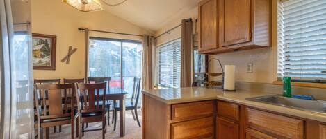 A kitchen with wooden cabinets, a dining area with chairs, a chandelier, and a window with blinds.