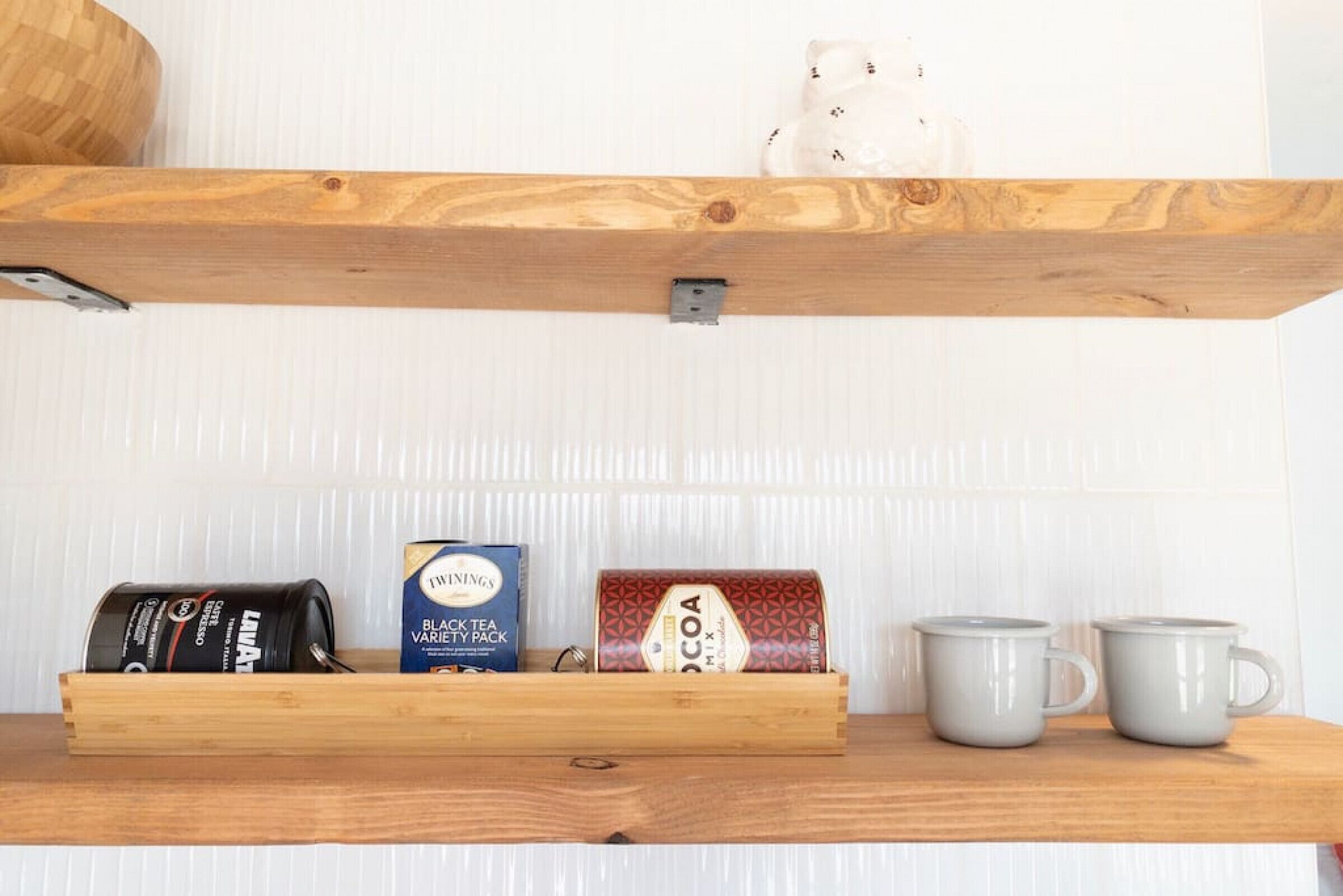 Une étagère en bois sur laquelle se trouvent une machine à café, une boîte contenant un assortiment de différents thés noirs, une boîte de chocolat en poudre et deux tasses grises.
