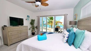 Habitación de hotel con cama, televisión, cómoda de madera, ventilador de techo, planta en maceta y vista a la playa.