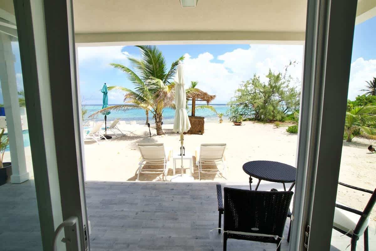 A beach view from a balcony with a table and chairs, palm trees, and a thatched umbrella.