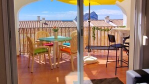 A room with a sliding glass door leading to an outdoor patio with a table and chairs, a yellow umbrella, and a view of the sea.