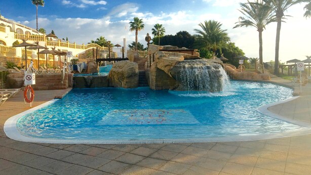 A swimming pool with a waterfall feature, surrounded by palm trees and a building with a balcony.