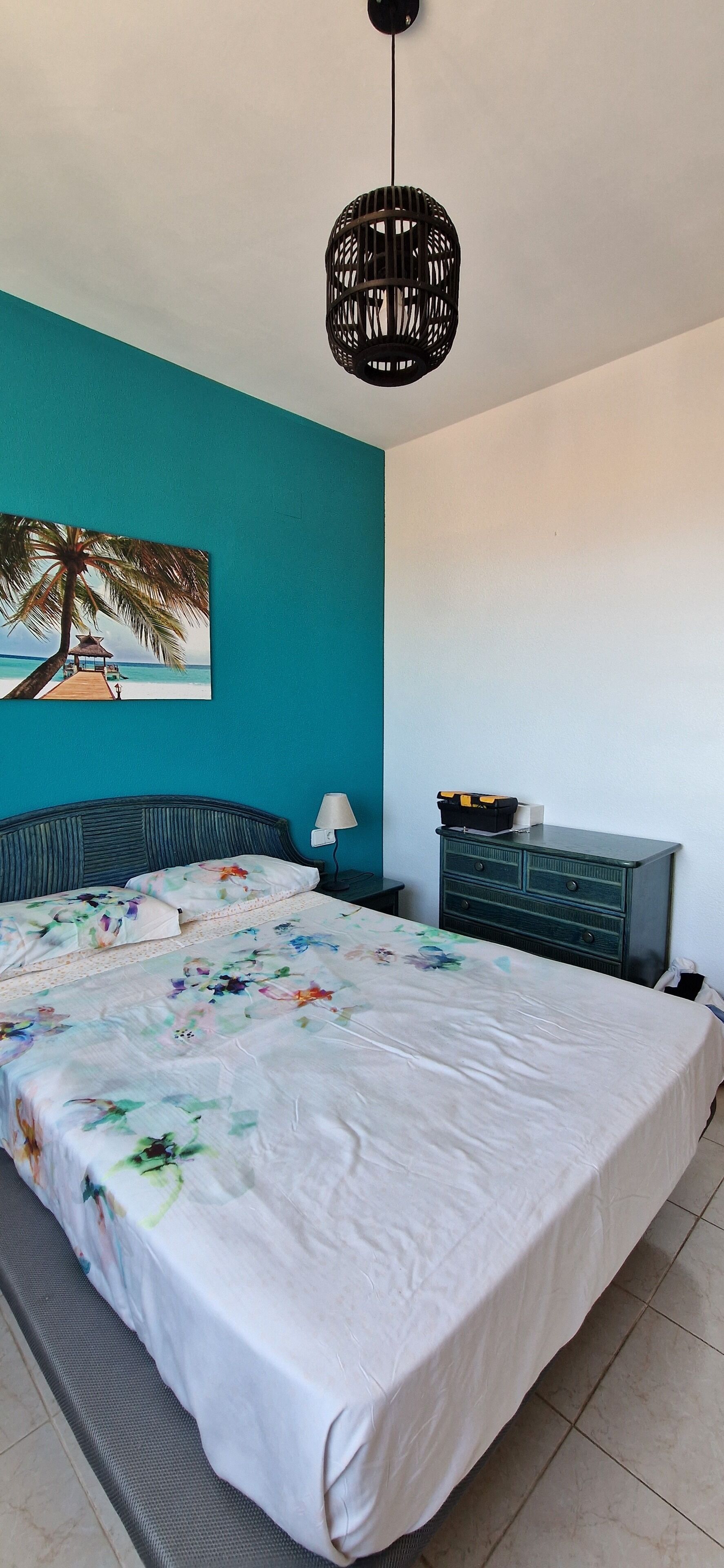Una camera da letto con un letto, una parete color petrolio, un quadro raffigurante una spiaggia, un comodino e un comò.