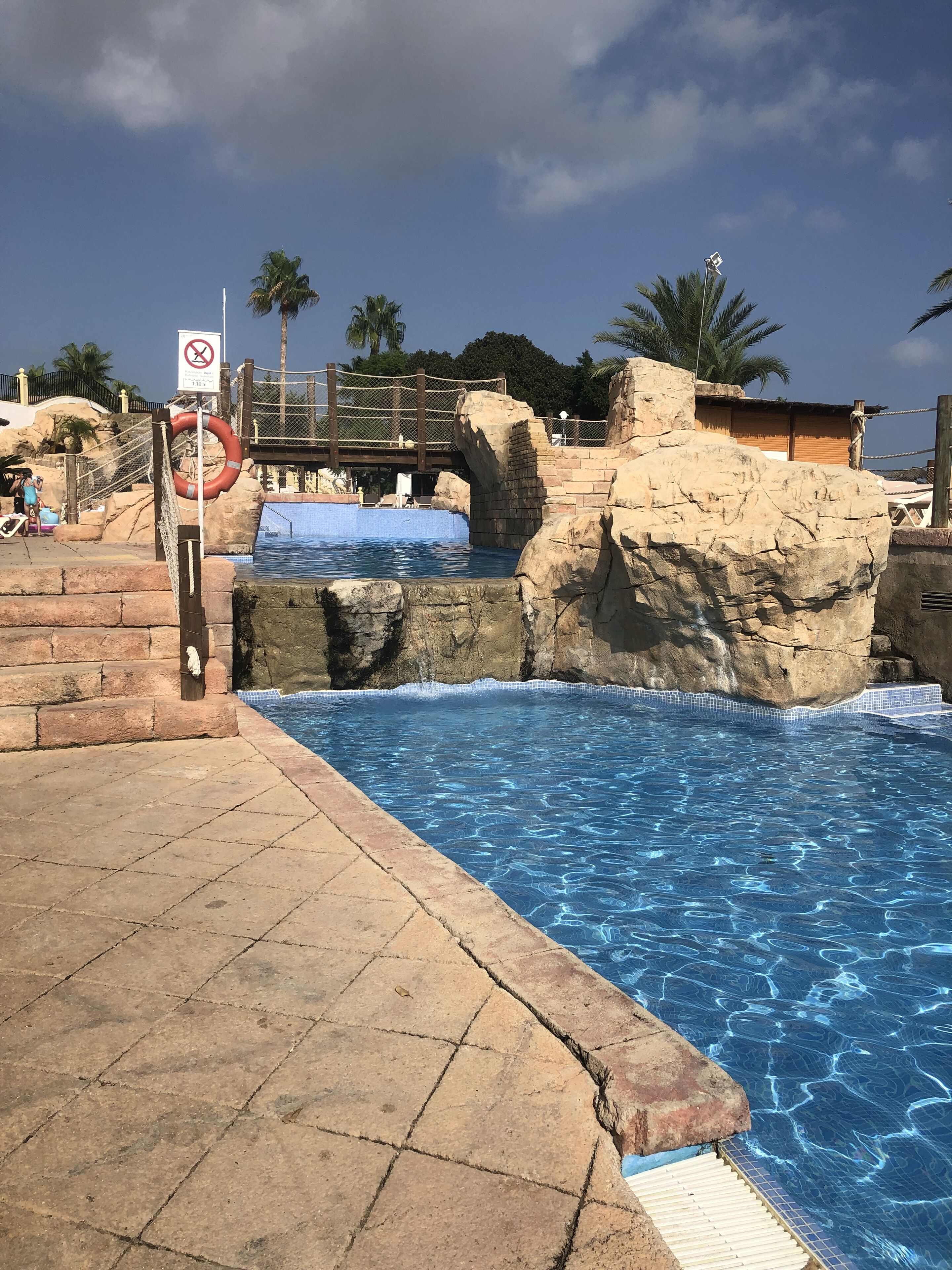 A swimming pool with a rocky edge, a no-entry sign, and a wooden bridge.