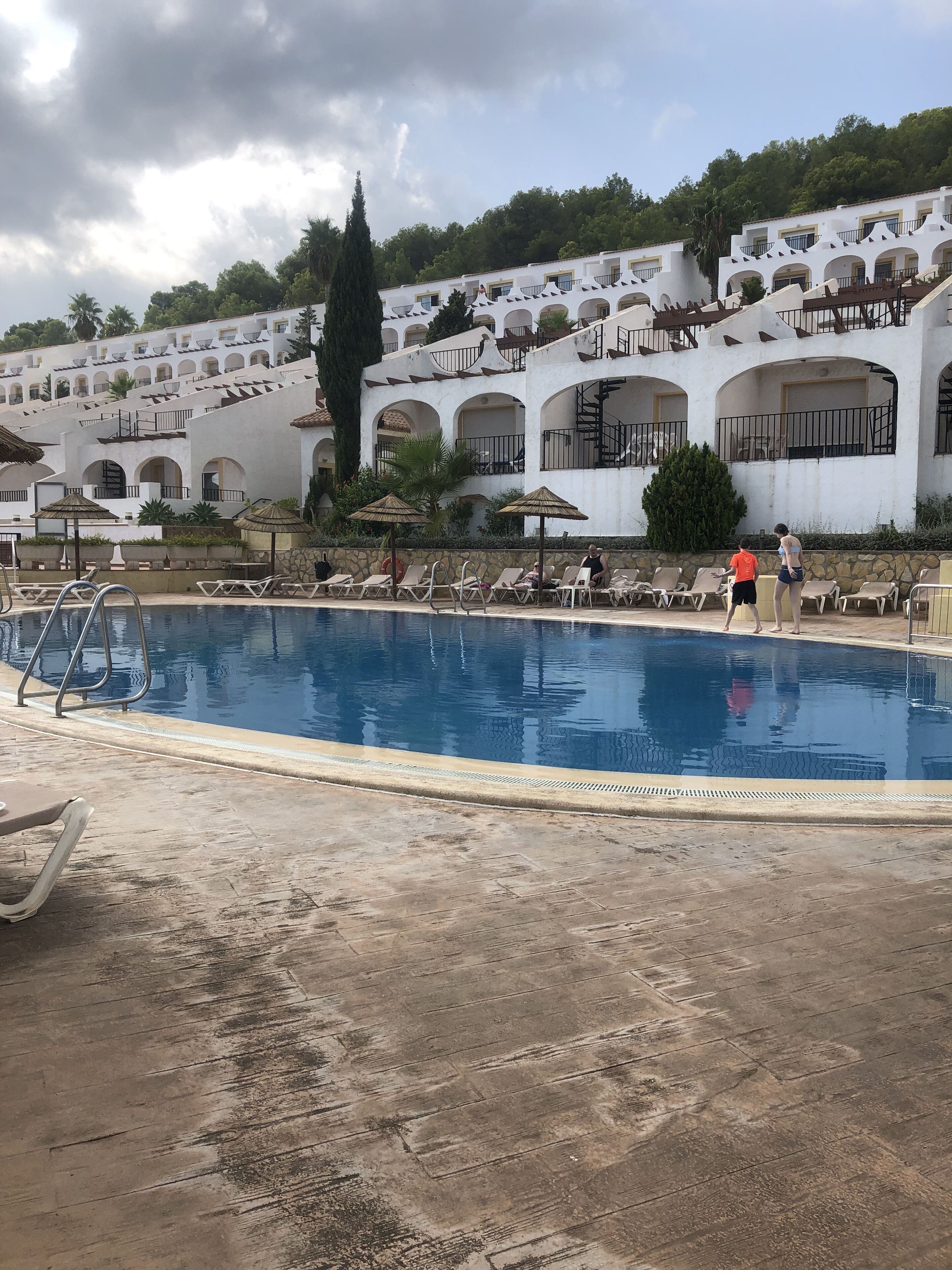 Una piscina con sillas de descanso y sombrillas, rodeada por un edificio blanco con arcos y balcones.