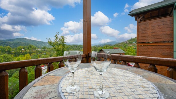 A table with two empty wine glasses on a tiled table, overlooking a scenic landscape with mountains and greenery. - Free Tickets | Million Dollar View, Easy Access, Arcade, Fireplace, Hot Tub (Gatlinburg)