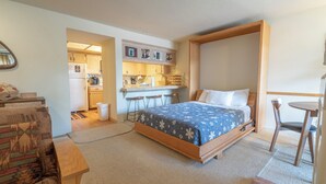 Ein Schlafzimmer mit Bett, kleinem Tisch, Stuhl und einer Küchenzeile im Hintergrund.