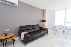 Un salon moderne avec un canapé en cuir noir, une table à manger en verre entourée de chaises, une petite table en bois sur laquelle est posée une plante, et un lampadaire.