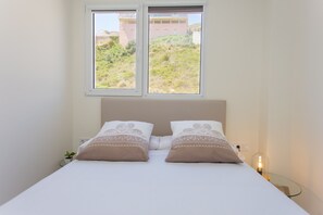 A bedroom with a bed, two windows, a bedside table with a lamp, and a view of greenery and buildings outside. - Cullera Dosel Beach Apartment (Cullera)