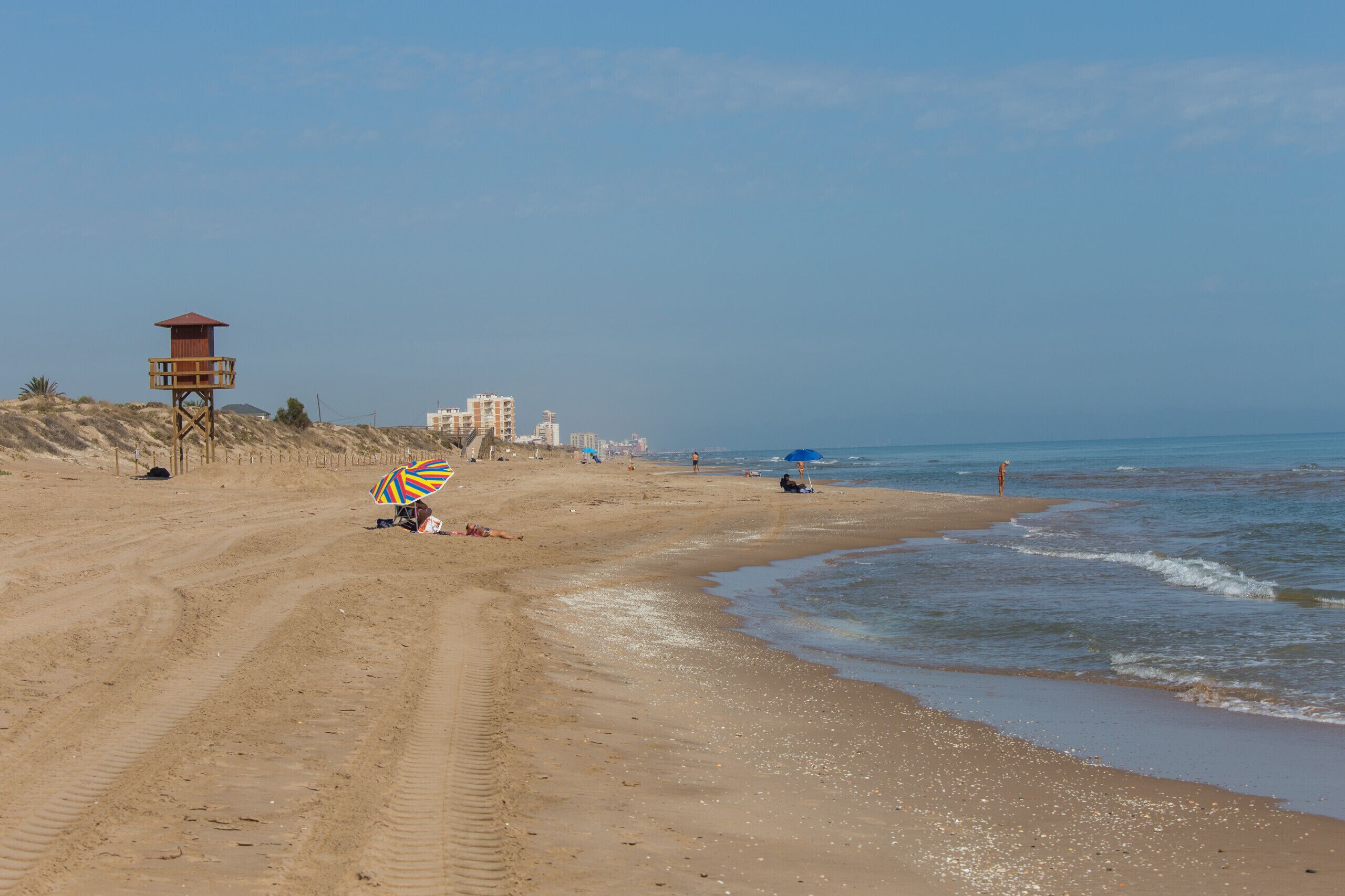 Une plage de sable avec un poste de surveillance pour les sauveteurs, quelques parasols et des bâtiments en arrière-plan.