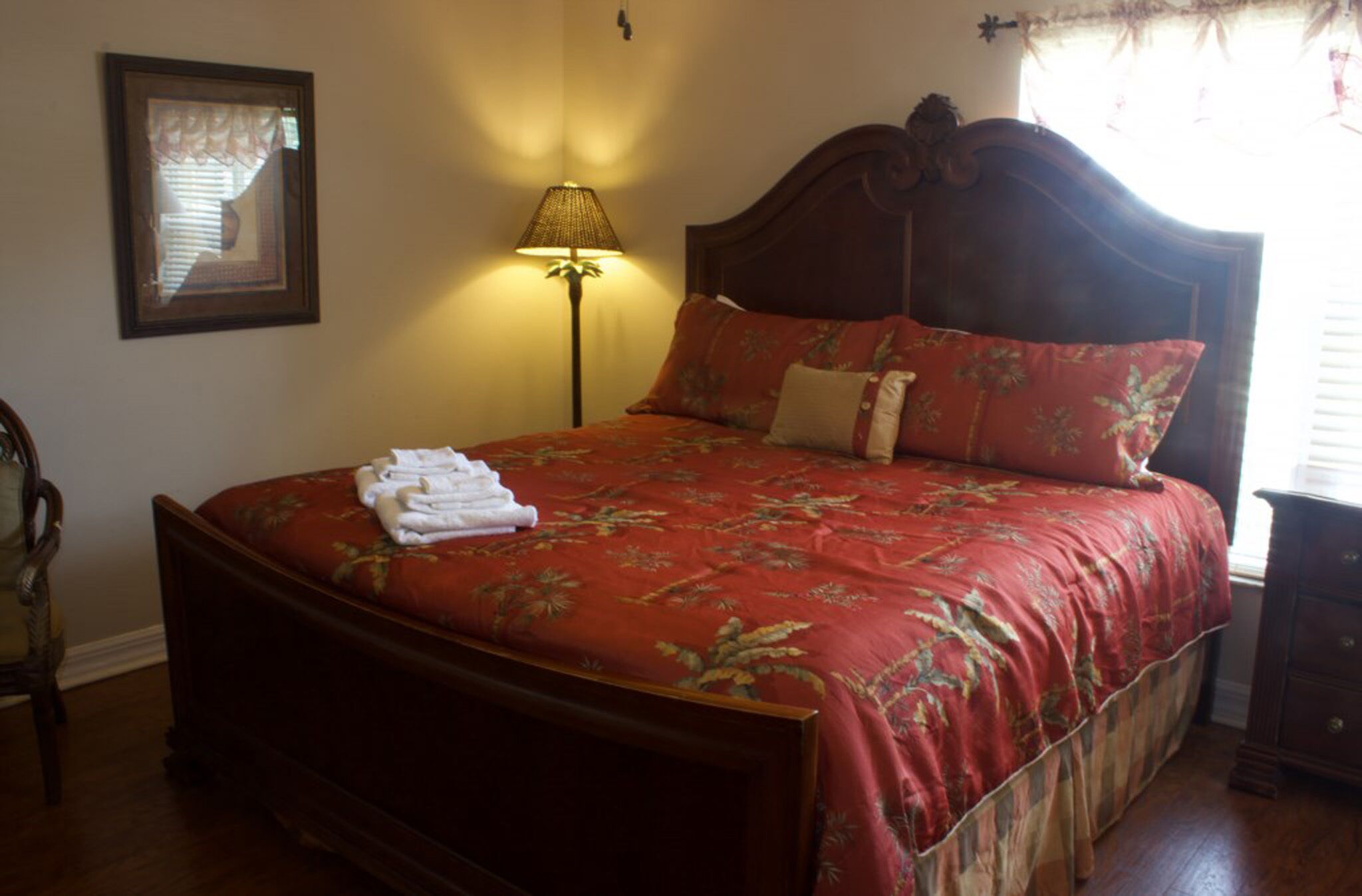 Una cama bien hecha con una colcha estampada con motivos florales, una lámpara de noche y un cuadro en la pared.