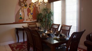 A dining room with a wooden table set for six, surrounded by chairs. A chandelier hangs above the table, and a large painting of animated characters is on the wall.