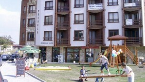 Un edificio de varios pisos con balcones y un área de juegos en la planta baja que incluye un sube y baja y columpios.