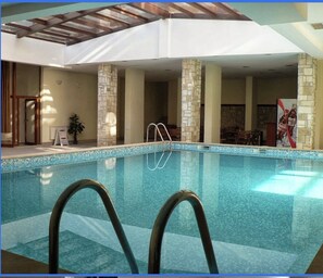 Ein beheiztes Hallenbad mit klarem Wasser, umgeben von steinernen Säulen und einer Holzbalkendecke.