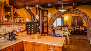 A rustic kitchen with wooden cabinets, a central island, hanging pendant lights, and a brick archway leading to a dining area. - House with private pool 90 km northern of Rome (montecampano)