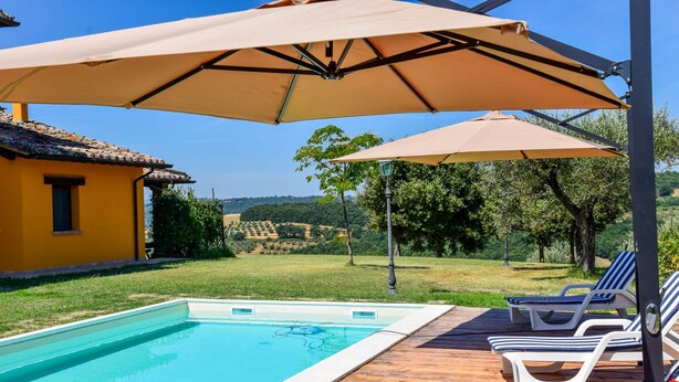 A swimming pool with lounge chairs and umbrellas, surrounded by a wooden deck and a yellow building. - House with private pool 90 km northern of Rome (montecampano)