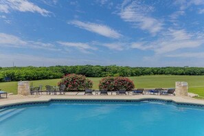 A swimming pool with lounge chairs and a stone wall, surrounded by greenery and a clear sky.
