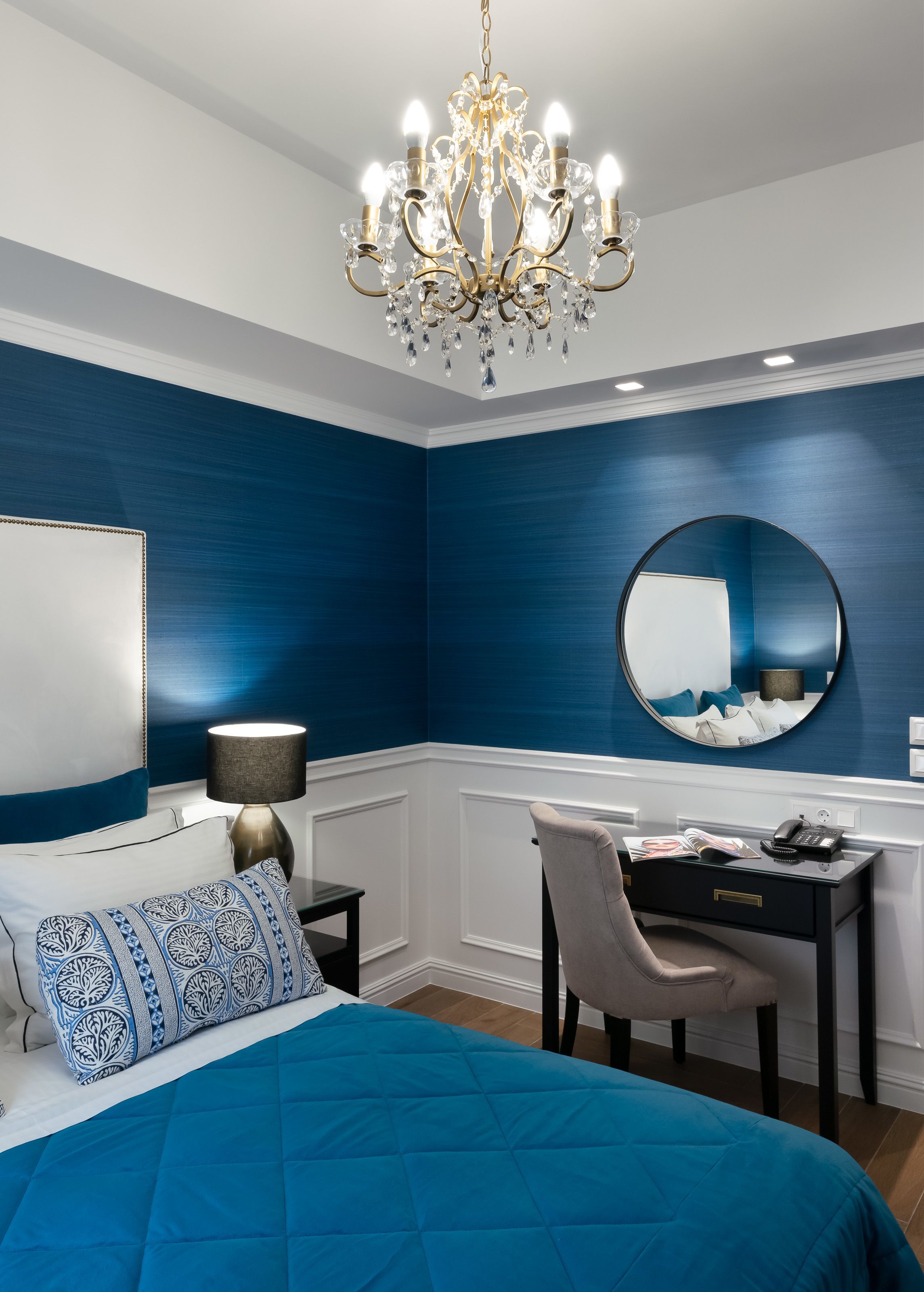 A bedroom with a bed, a desk, a chair, a lamp, a mirror, and a chandelier.