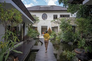 A pathway leading to a building with a circular window, surrounded by greenery and a small water feature. - The ultimate Bali beachside getaway at Villa Kaira Estate, a 7-bedroom luxury villa in Berawa, Canggu. The Estate offers daily breakfast, a full-time private chef, airport transfers, complimentary massages, housekeeping, and a dedicated driver. (North Kuta)