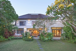 A modern house with a well-maintained garden, a patio with seating, and a large window overlooking the garden. - The ultimate Bali beachside getaway at Villa Kaira Estate, a 7-bedroom luxury villa in Berawa, Canggu. The Estate offers daily breakfast, a full-time private chef, airport transfers, complimentary massages, housekeeping, and a dedicated driver. (North Kuta)