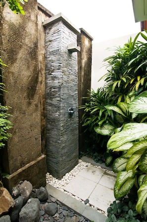 A stone shower enclosure with a metal handle, surrounded by greenery and pebbles.