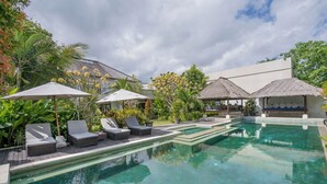 A pool area with lounge chairs and umbrellas, surrounded by lush greenery and a thatched-roof structure. - The ultimate Bali beachside getaway at Villa Kaira Estate, a 7-bedroom luxury villa in Berawa, Canggu. The Estate offers daily breakfast, a full-time private chef, airport transfers, complimentary massages, housekeeping, and a dedicated driver. (North Kuta)