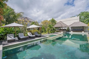 A pool area with lounge chairs and umbrellas, surrounded by lush greenery and a thatched-roof structure.