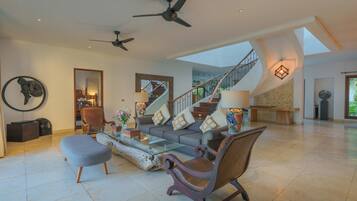 A spacious living room with a modern sofa, armchairs, and a glass coffee table. A staircase with a railing leads to an upper level.