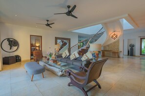 A spacious living room with a modern sofa, armchairs, and a glass coffee table. A staircase with a railing leads to an upper level.