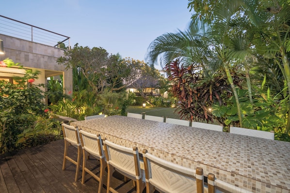 A covered outdoor dining area with a tiled table and chairs, surrounded by lush greenery and a view of a building in the background.