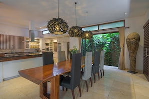 A modern dining area with a wooden table and chairs, pendant lights, and a view of greenery outside. - The ultimate Bali beachside getaway at Villa Kaira Estate, a 7-bedroom luxury villa in Berawa, Canggu. The Estate offers daily breakfast, a full-time private chef, airport transfers, complimentary massages, housekeeping, and a dedicated driver. (North Kuta)