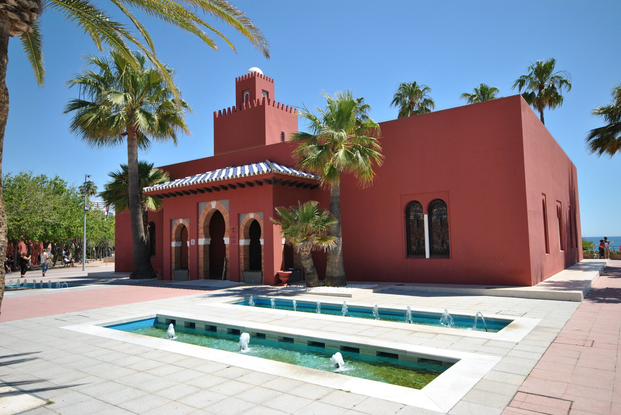 Ein rotes Gebäude mit Rundbogenfenstern und einem Springbrunnen davor.