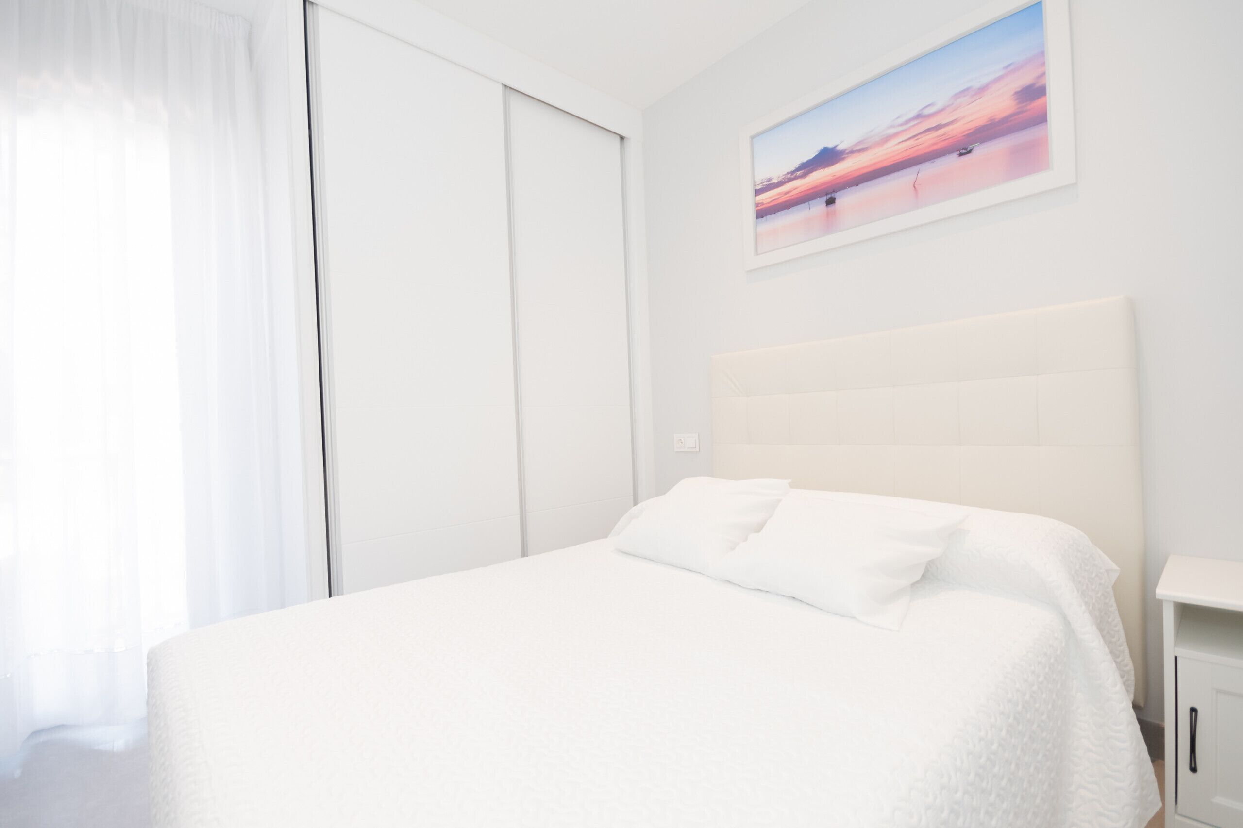 A white bed with a headboard, a white nightstand, and a painting of a sunset on the wall.