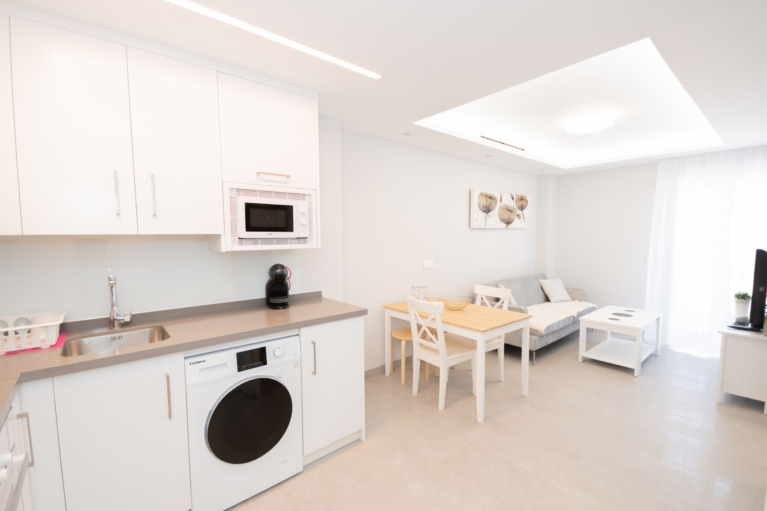 A modern kitchen with a dishwasher, microwave, and coffee maker. There is a dining area with a table and chairs, and a sofa.