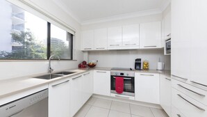 A modern kitchen with white cabinets, a Bosch dishwasher, a red oven mitt, and a window with a view of buildings and trees. - Newly Renovated Unit near Forster Main Beach! (Forster)