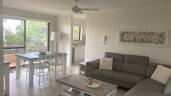 Ein modernes Wohnzimmer mit grauem Sofa, einem Esstisch mit Stühlen, einem Couchtisch, einem Deckenventilator und einer Küche im Hintergrund.