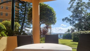 A patio area with a round table and two chairs, surrounded by greenery and a view of a building and hills in the distance.