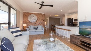 Una sala moderna con una pared de ladrillo decorativa, un ventilador de techo y un televisor grande que muestra una escena de playa.