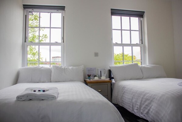 A hotel room with two single beds, a nightstand with a lamp, and two windows with a view of greenery and buildings. - Crofts Hotel (Cardiff)