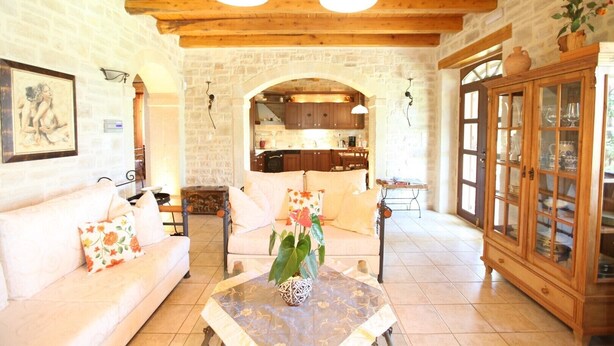 A cozy living room with a stone wall, wooden ceiling beams, a sofa, a coffee table, and a cabinet with glass doors. - Villa Persephone, drofnah and beach (Rethymnon)