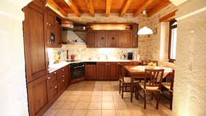 A kitchen with wooden cabinets, a built-in oven, a dining table with chairs, and a stone wall. - Villa Persephone, drofnah and beach (Rethymnon)
